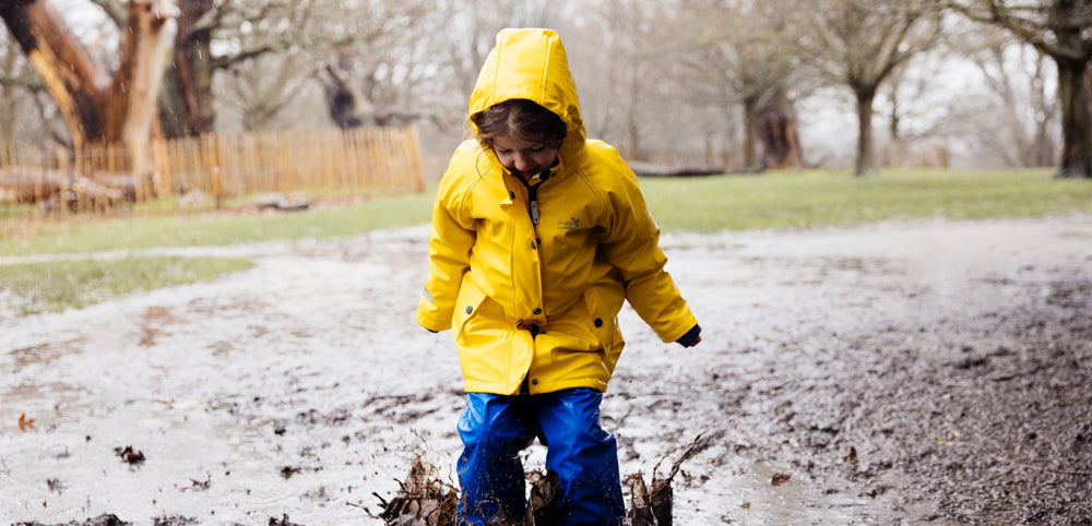 girl jumping into muddy puddle wearing yellow waterproof jacket with hood up, and blue waterproof trousers