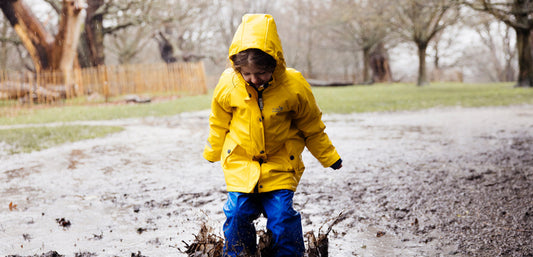 girl jumping into muddy puddle wearing yellow waterproof jacket with hood up, and blue waterproof trousers