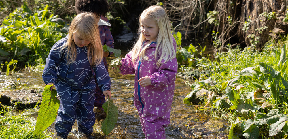 girls paddling in shallow water holding leaves, wearing colourful muddy puddles waterproof puddle suits
