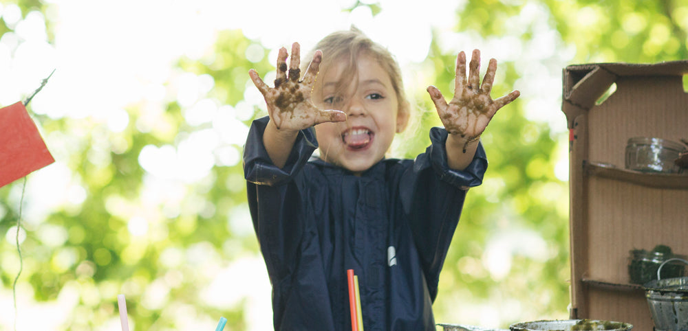 girl at forest school wearing navy puddle suit and holding up muddy hands