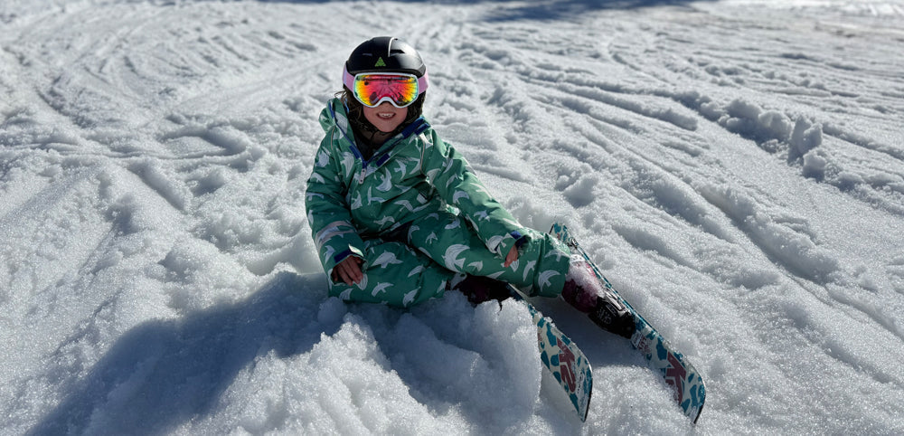 child sitting in snow wearing green bird print skiwear, ski goggles, helmet and skis