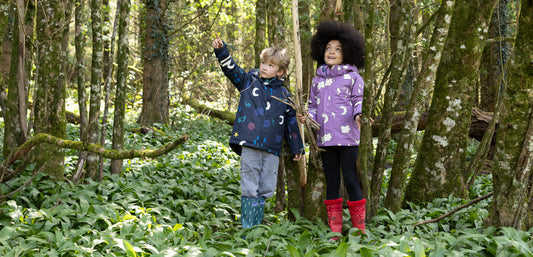 boy and girl standing in woodland looking up at trees, wearing vibrant muddy puddles winter jackets and wellies