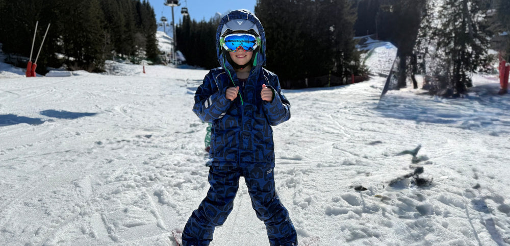 child on ski slope wearing muddy puddles blue ski outfit, ski goggles, helmet and skis
