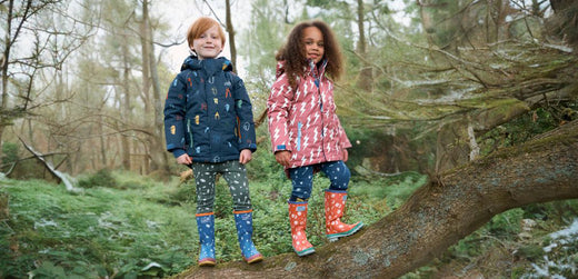 children standing on a tree branch wearing colourful muddy puddles ski jackets and wellies