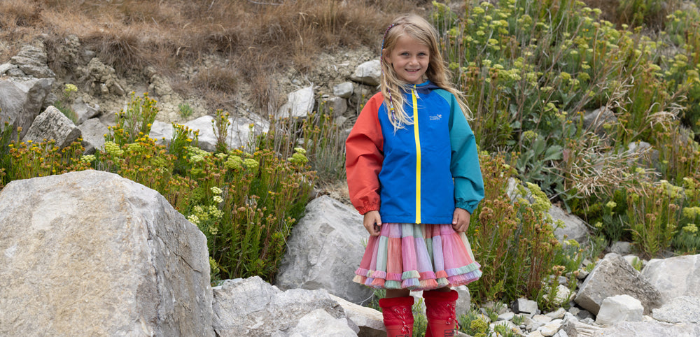 girl stood by rocky hillside wearing vibrant colourblock raincoat by muddy puddles, tutu skirt and red wellies
