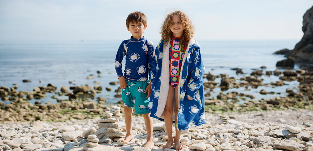 Two children in colorful swimwear and blue sun-print cover-ups standing on a rocky beach