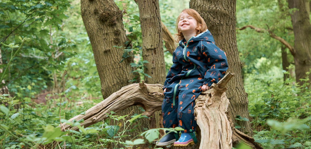 smiling child sitting on tree branch, looking up and wearing muddy puddles navy puddlesuit and wellies