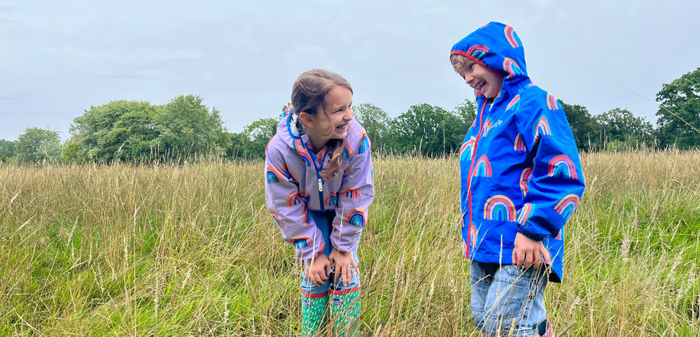 Two children in colorful waterproof jackets with rainbow patterns playing outdoors in a grassy field.