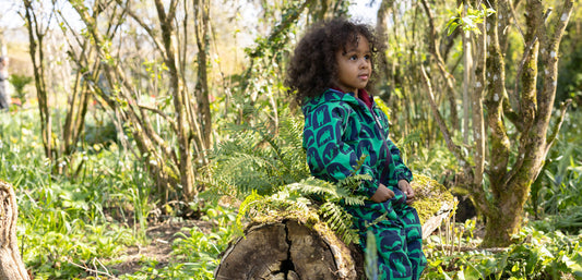 boy sitting on a log in green woodland area, wearing muddy puddles cave print puddlesuit