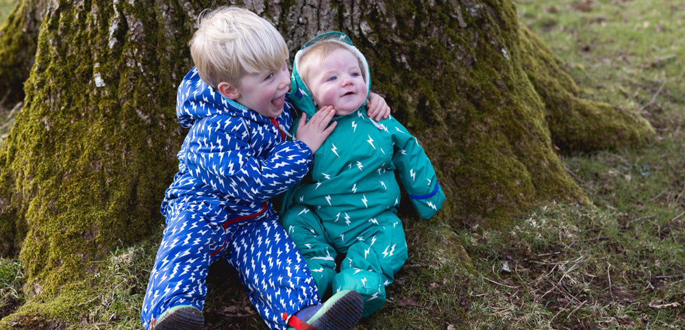 toddler and baby sat at bottom of moss covered tree wearing blue lightning puddlesuit and green lightning puddlesuit