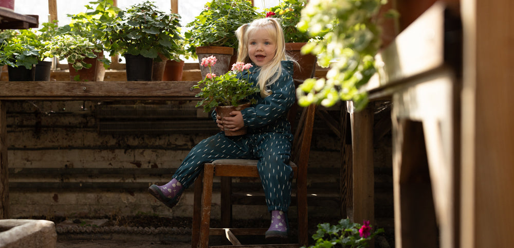 blonde girl sitting on a chair in a greenhouse holding a plant, wearing green muddy puddles waterproof puddlesuit