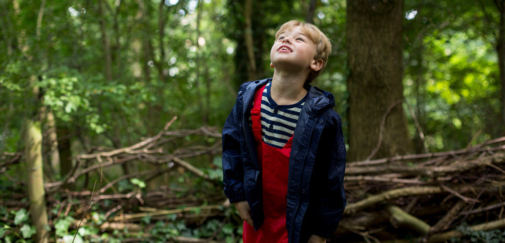 Young boy in durable, waterproof red overalls and rain jacket, looking upward in a dense forest, embodying Muddy Puddles' ethos of outdoor exploration.