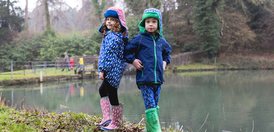 boy and girl stood by lake, wearing vibrant waterproof jackets, trapper hats and wellies