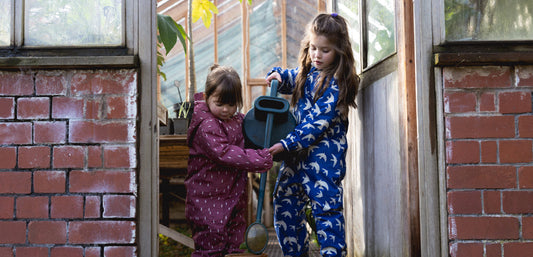 two girls stood at entrance to greenhouse holding a watering can and wearing muddy puddles pink raindrop scampsuit and blue bird puddlesuit