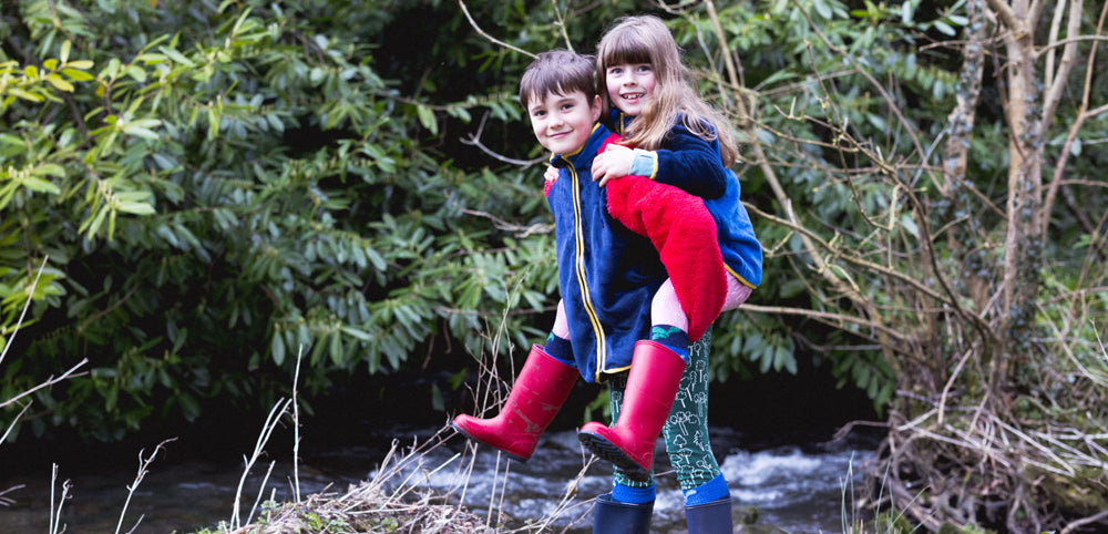 Two children in waterproof jackets and red wellies by a stream in a lush outdoor setting