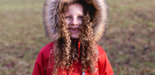 Child outdoors wearing a red waterproof jacket with fur-lined hood, curly hair visible