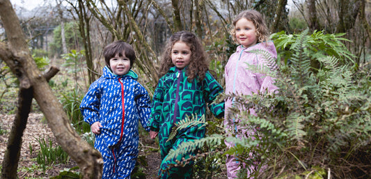three children holding hands in woodland area wearing blue lightning puddlesuit, green cave puddlesuit, and pink forest puddlesuit