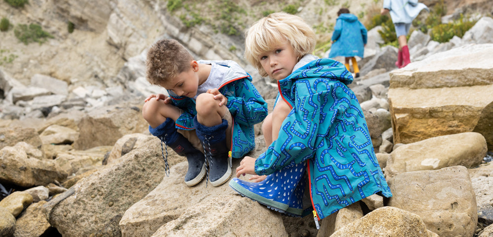 two boys on rocky beach wearing muddy puddles waterproof jackets and wellies