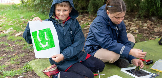 Children in waterproof jackets doing outdoor crafts on grass, holding art print