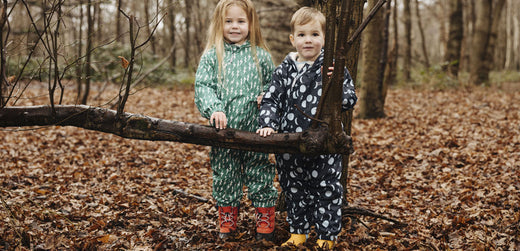 Two children in waterproof rain suits and boots standing in a leaf-covered forest