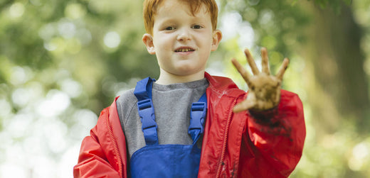 Child outdoors in waterproof jacket and dungarees, showing muddy hand, kids rainwear