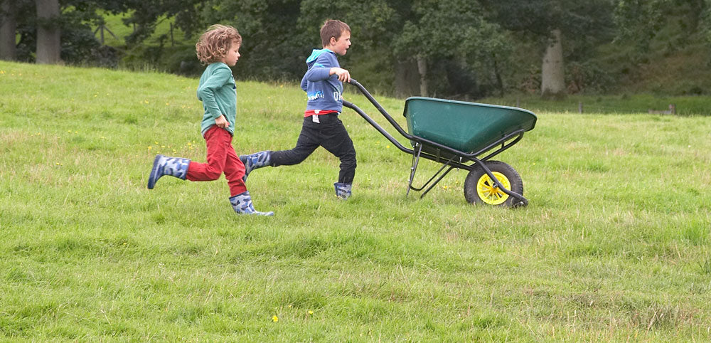 Children in waterproof boots running outdoors with a wheelbarrow on grassy field