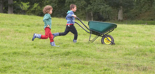Children in waterproof boots running outdoors with a wheelbarrow on grassy field