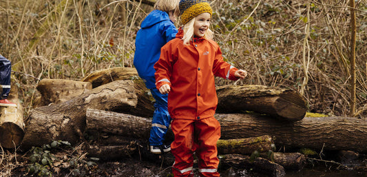 boy and girl standing in front of logs in woodland area wearing red and blue muddy puddles puddlesuits