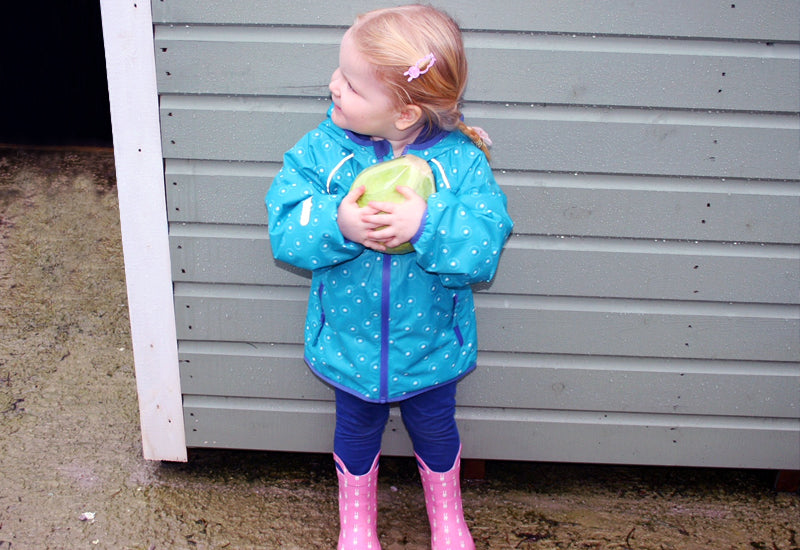 toddler girl stood in front of a shed holding a lettuce, wearing vibrant blue muddy puddles waterproof jacket