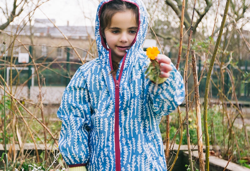 girl stood in garden holding a flower, wearing vibrant blue muddy puddles raincoat