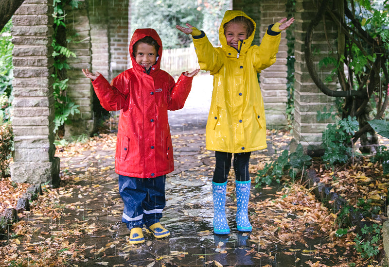 Two children in waterproof raincoats and boots enjoy the rain under a leafy archway, highlighting Muddy Puddles' durable, outdoor clothing for kids.
