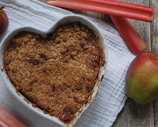close up of apple and rhubarb crumble in a heart shaped dish, with apples and rhubarb in the background