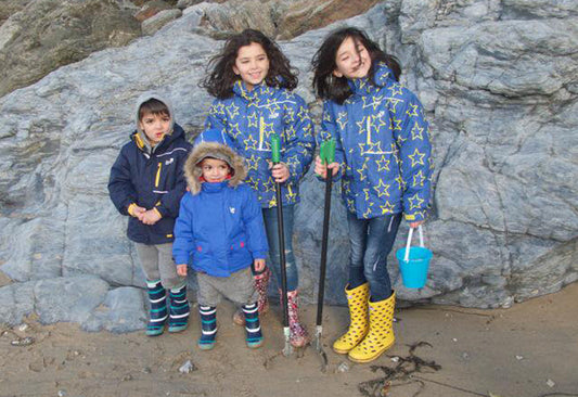 children standing on sandy beach, wearing muddy puddles waterproof jackets and wellies
