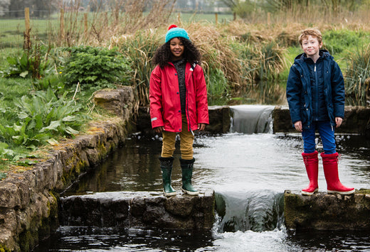 boy and girl standing in a stream wearing classic red and navy muddy puddles raincoats and wellies