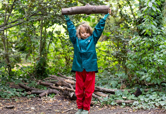 a boy holding a log above his head in a woodland area, wearing a muddy puddles waterproofs