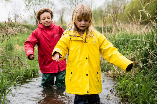 children wading through a deep puddle, wearing bright yellow and red muddy puddles waterproof jackets