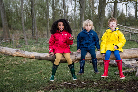 children sitting on a tree branch, wearing bold muddy puddles red navy and yellow raincoats and wellies