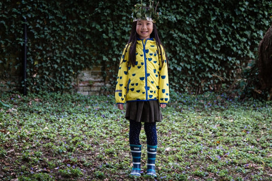 girl stood in front of an ivy covered wall, wearing vibrant yellow waterproof jacket, striped wellies and a crown made from leaves