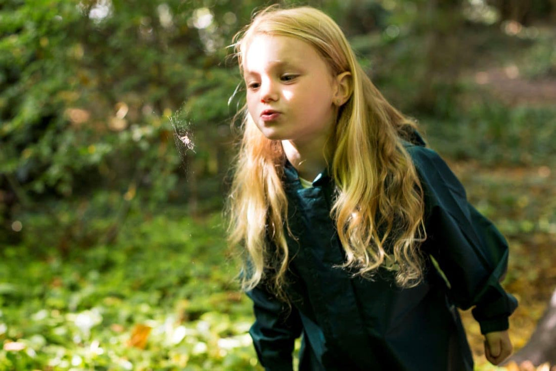 girl with long blonde hair blowing air on a spiders web, wearing green muddy puddles puddlesuit