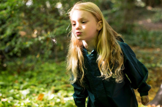 girl with long blonde hair blowing air on a spiders web, wearing green muddy puddles puddlesuit