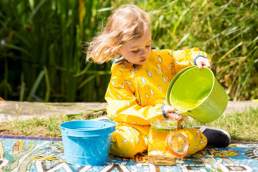 girl sitting on picnic blanket pouring water into a jar, wearing a bright yellow muddy puddles puddlesuit