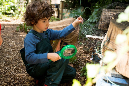 boy at forest school wearing green waterproof dungarees, holding a magnifying glass