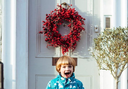 child stood in front of a door smiling, with a red berry wreath hanging on the door