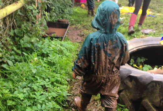child walking through forest school area, with their back to the camera, wearing a green puddlesuit covered in mud