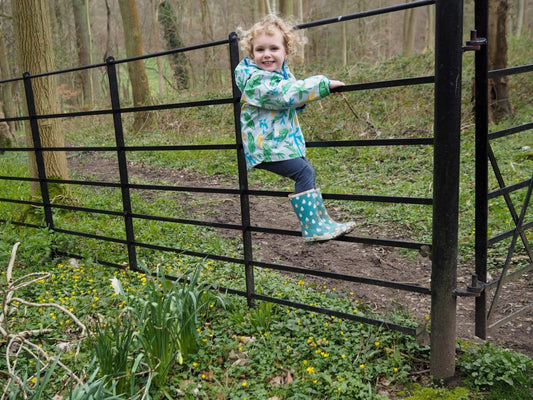 toddler climbing on a black woodland fence, wearing green muddy puddles waterproof jacket and wellies
