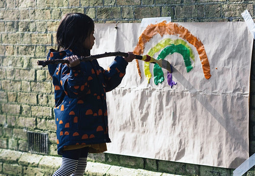 child painting a rainbow on a sheet of paper stuck to a brick wall, using a paintbrush attached to a long stick, wearing a navy muddy puddles raincoat