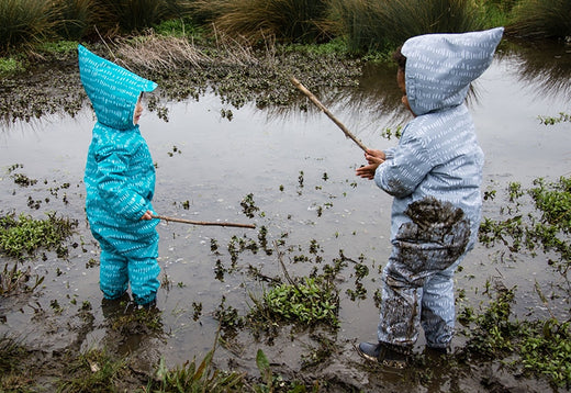 toddler children standing in a muddy puddle holding sticks, wearing blue and green muddy puddles waterproof scampsuits