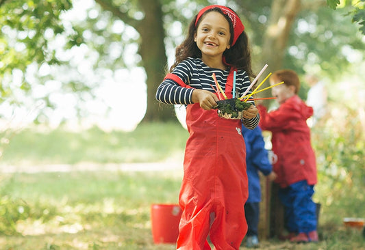girl attending forest school, holding a mud pie, wearing red muddy puddles waterproof dungarees and bandana