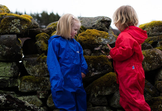 children stood in front of mossy wall, wearing red and blue muddy puddles puddlesuits