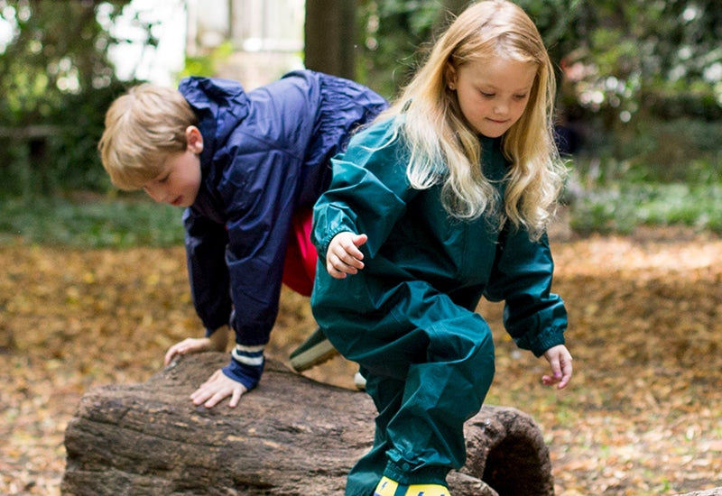 girl and boy climbing on log, in woodland area, wearing muddy puddles waterproofs